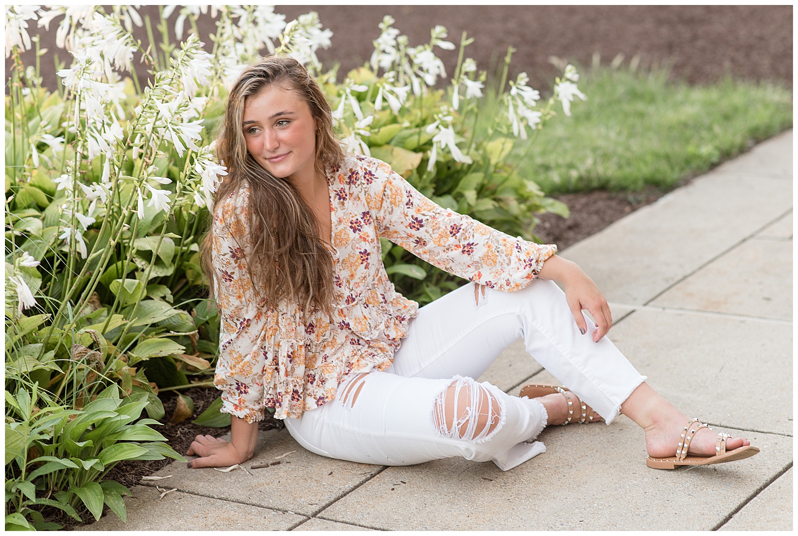 senior girl sitting on pathway beside blooming hosta plants looking over right shoulder with left arm resting on bent left leg