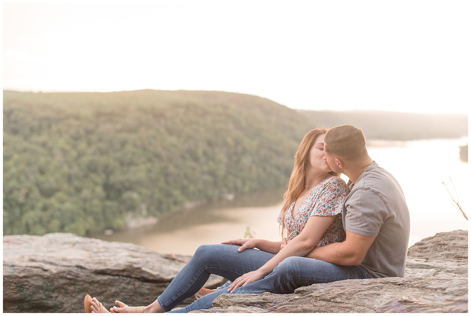 engaged couple sitting closely on large rock kissing atop pinnacle overlook with susquehanna river behind them