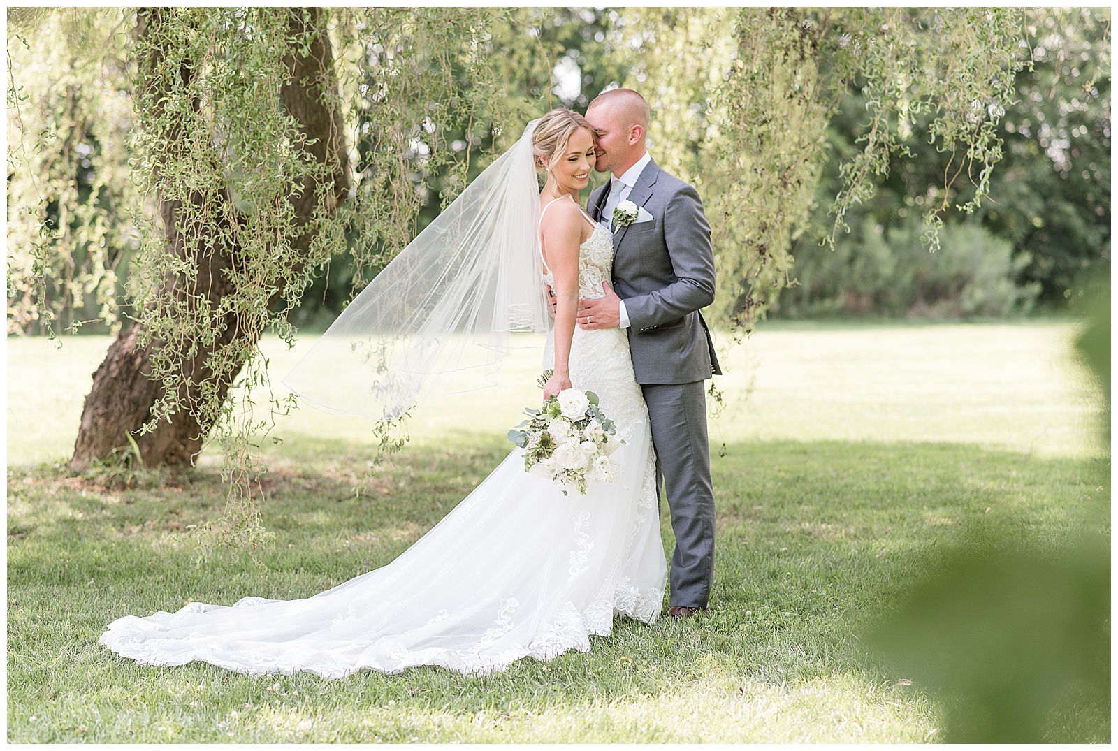 groom hugs and kisses bride as she holds bouquet in right hand standing under willow tree in lancaster pennsylvania