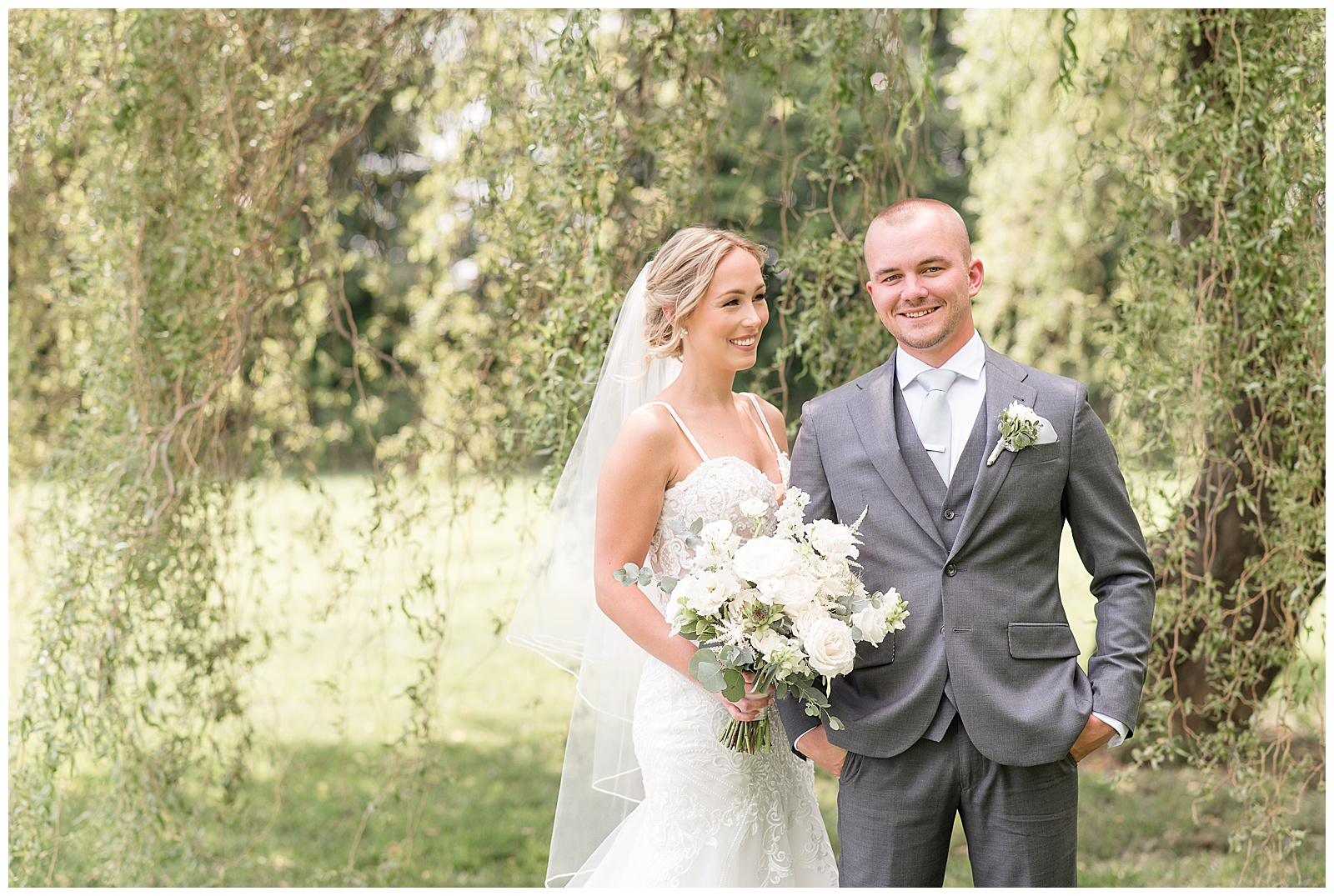 bride smiles as she looks at her groom who is smiling at the camera under willow tree at barn at silverstone