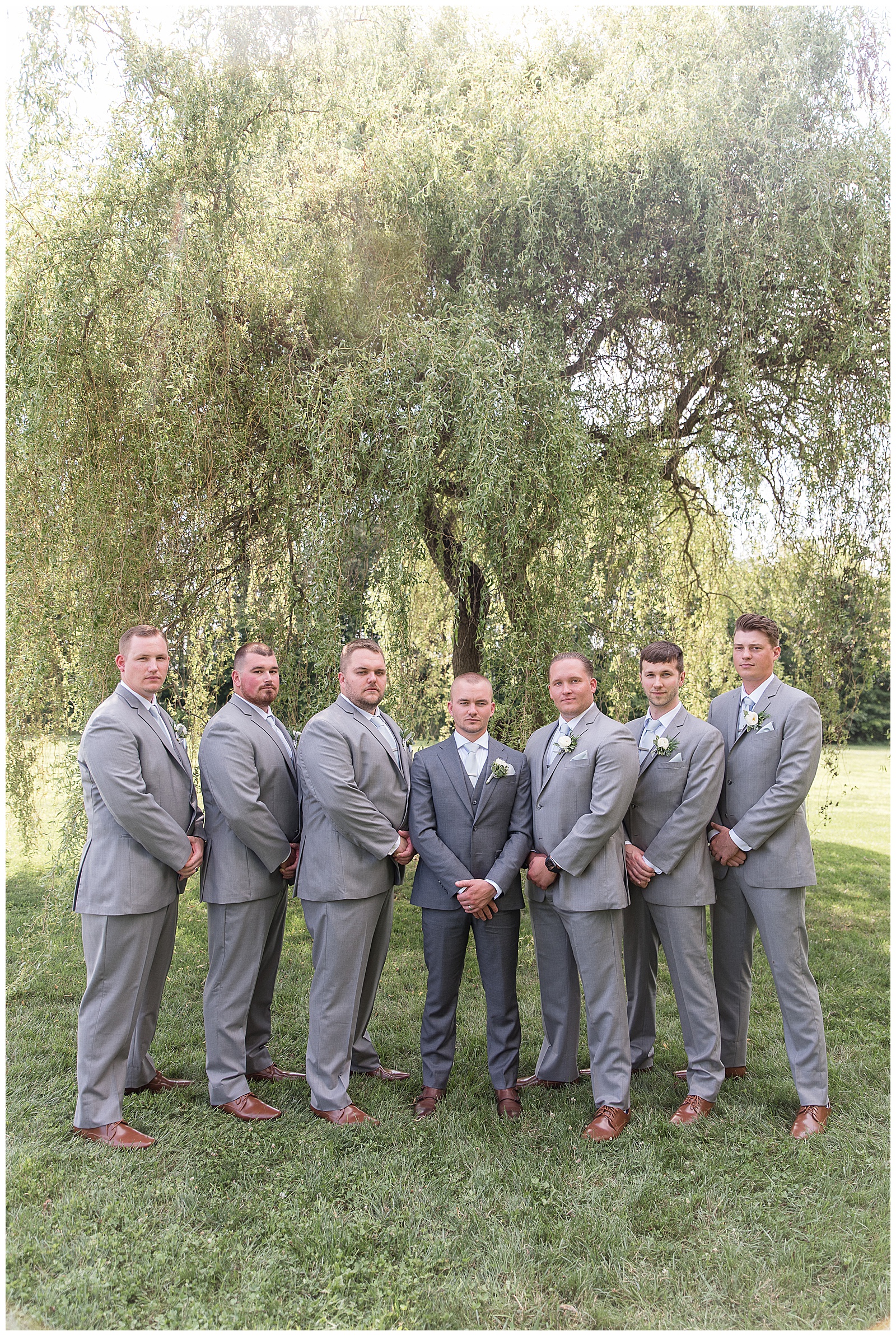groom surrounded by six groomsmen all in gray suits with hands folded in front of them under willow tree in lancaster pennsylvania