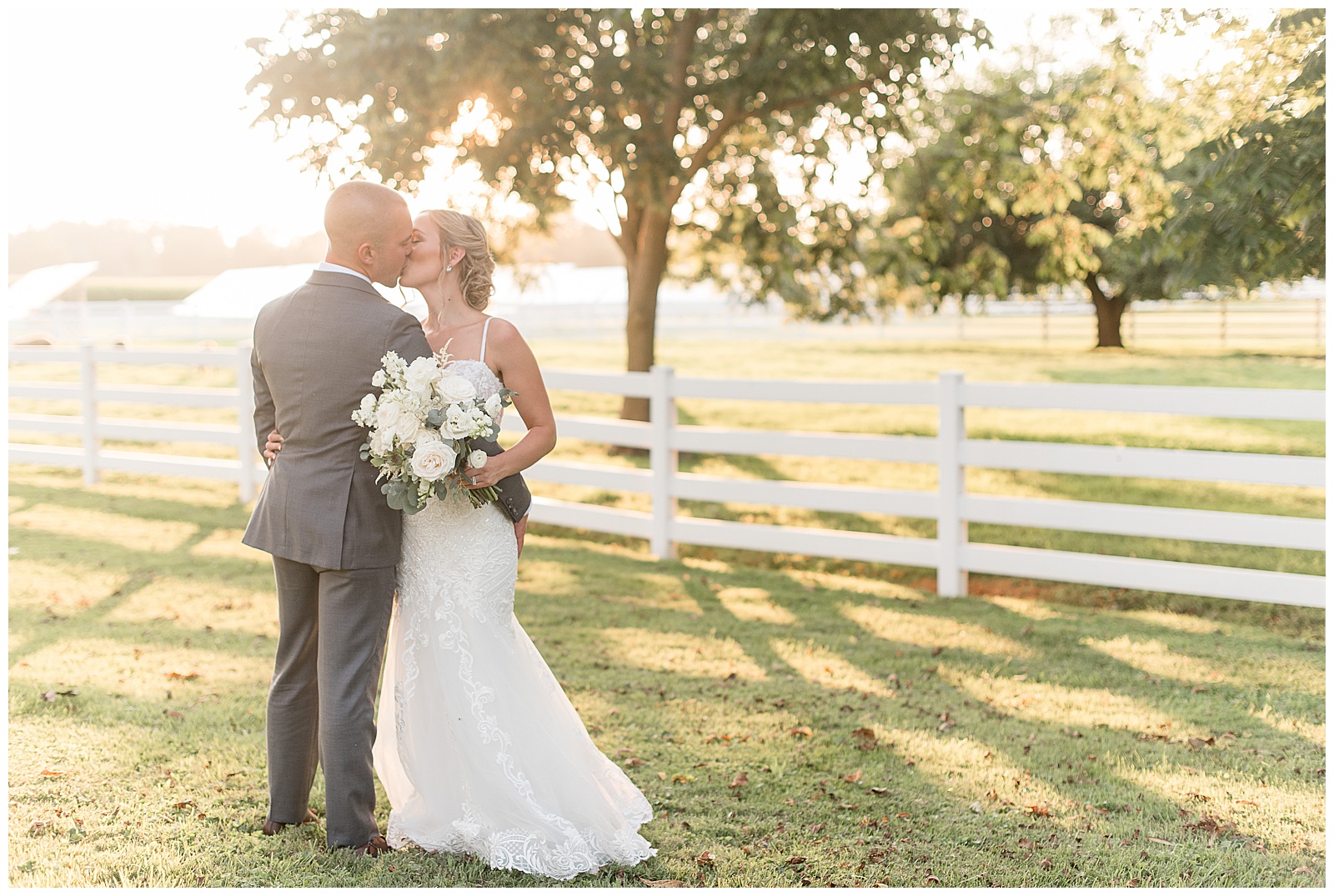 bride and groom kiss and hug beside white fence on sunny evening at the barn at silverstone in lancaster pennsylvania