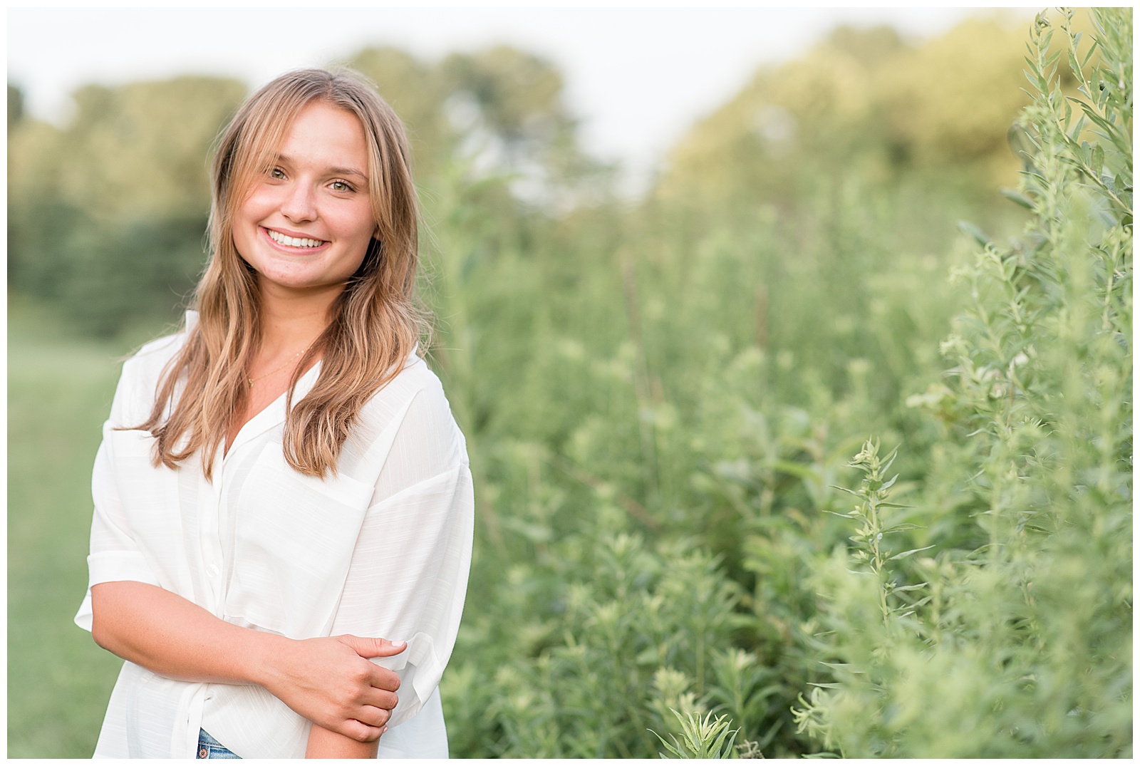close up of senior girl next to tall grasses with right hand holding left elbow on sunny evening in lancaster, pennsylvania
