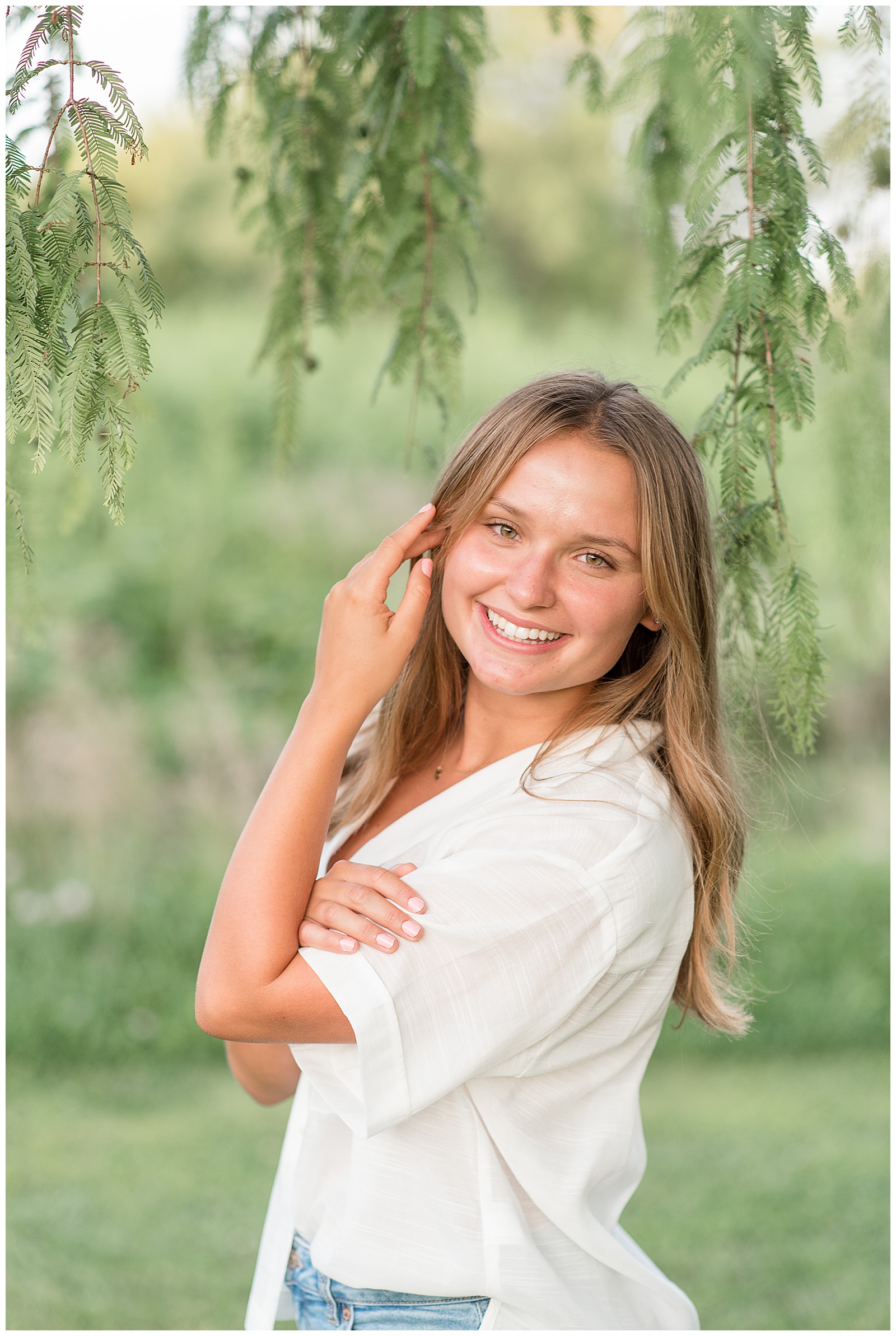 senior girl using left hand to keep her long brown hair out of her face as she smiles at camera under tree at lancaster county park