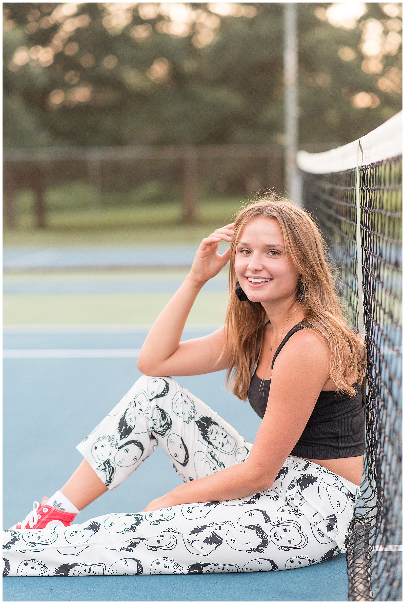 senior girl leaning at tennis court net with right elbow on bent right knee at lancaster county park