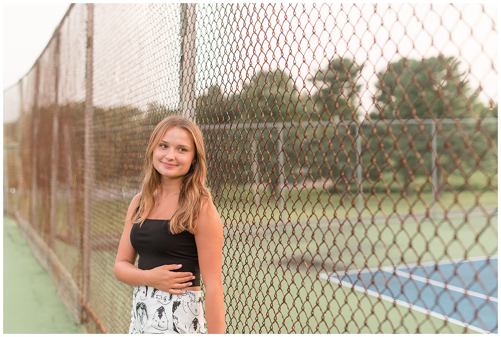 senior girl leaning against chain link fence of tennis courts with right hand across stomach at lancaster county park