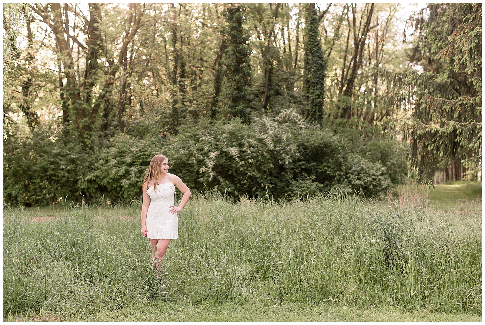 senior girl in white spaghetti strap dress with left hand on hip standing among tall wild grasses at overlook park in lancaster pennsylvania