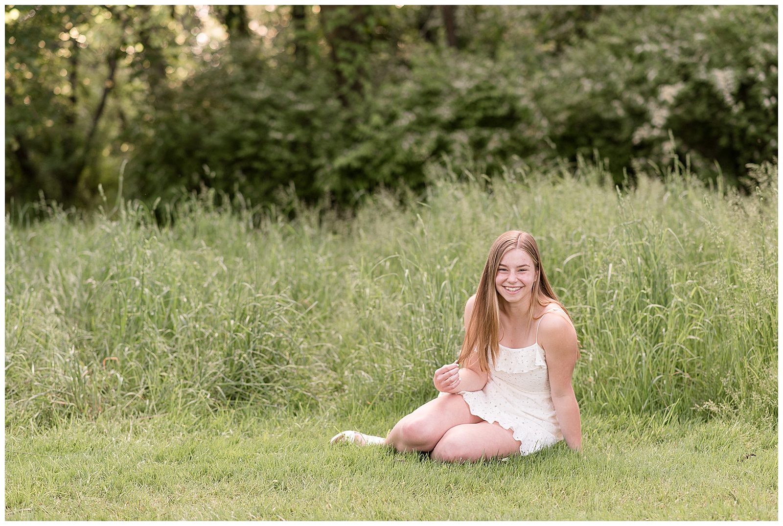 senior girl sitting on the grass by tall grasses smiling at camera on sunny evening at overlook park