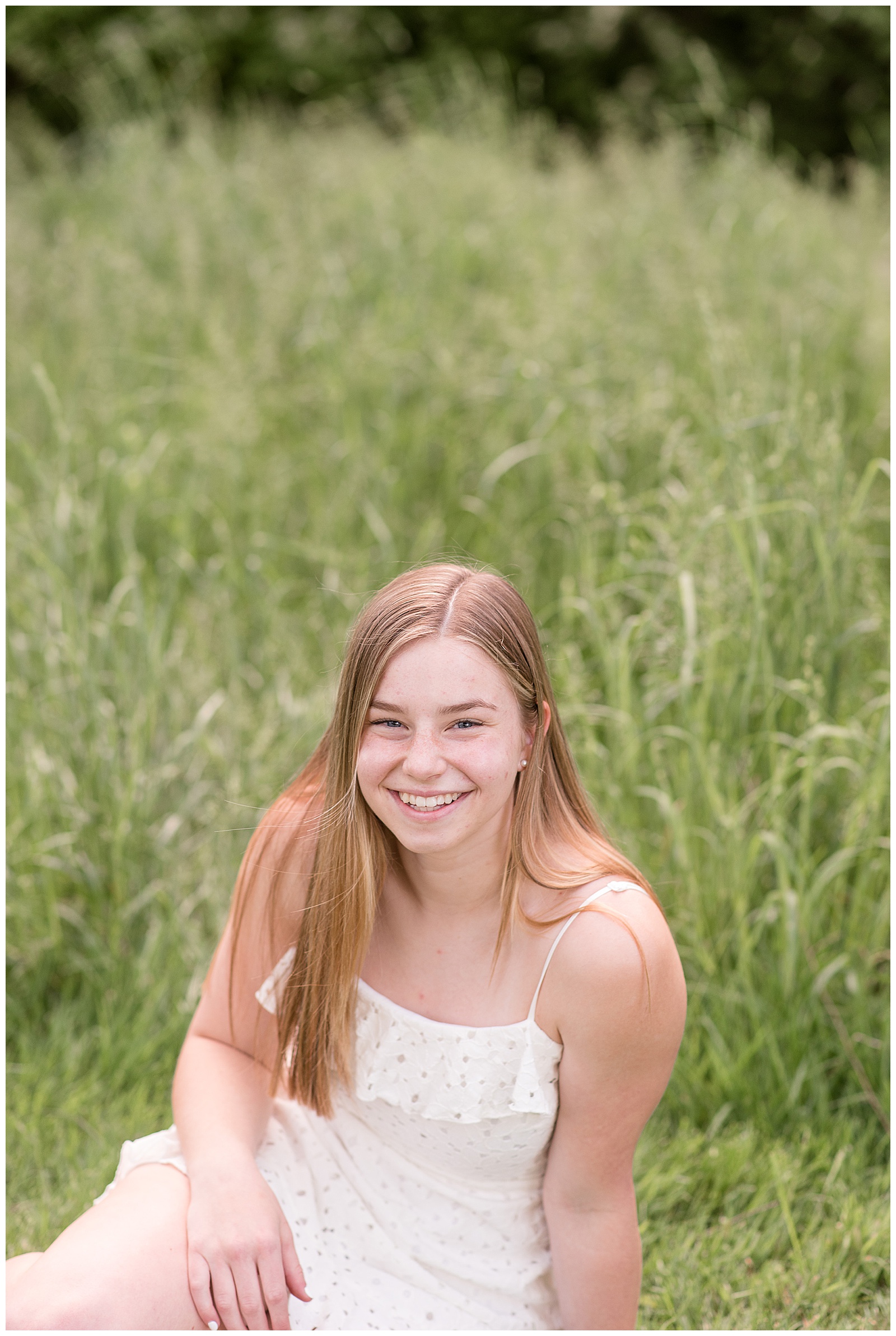 closeup photo of senior girl looking up at camera smiling as she sits by tall wild grasses at overlook park