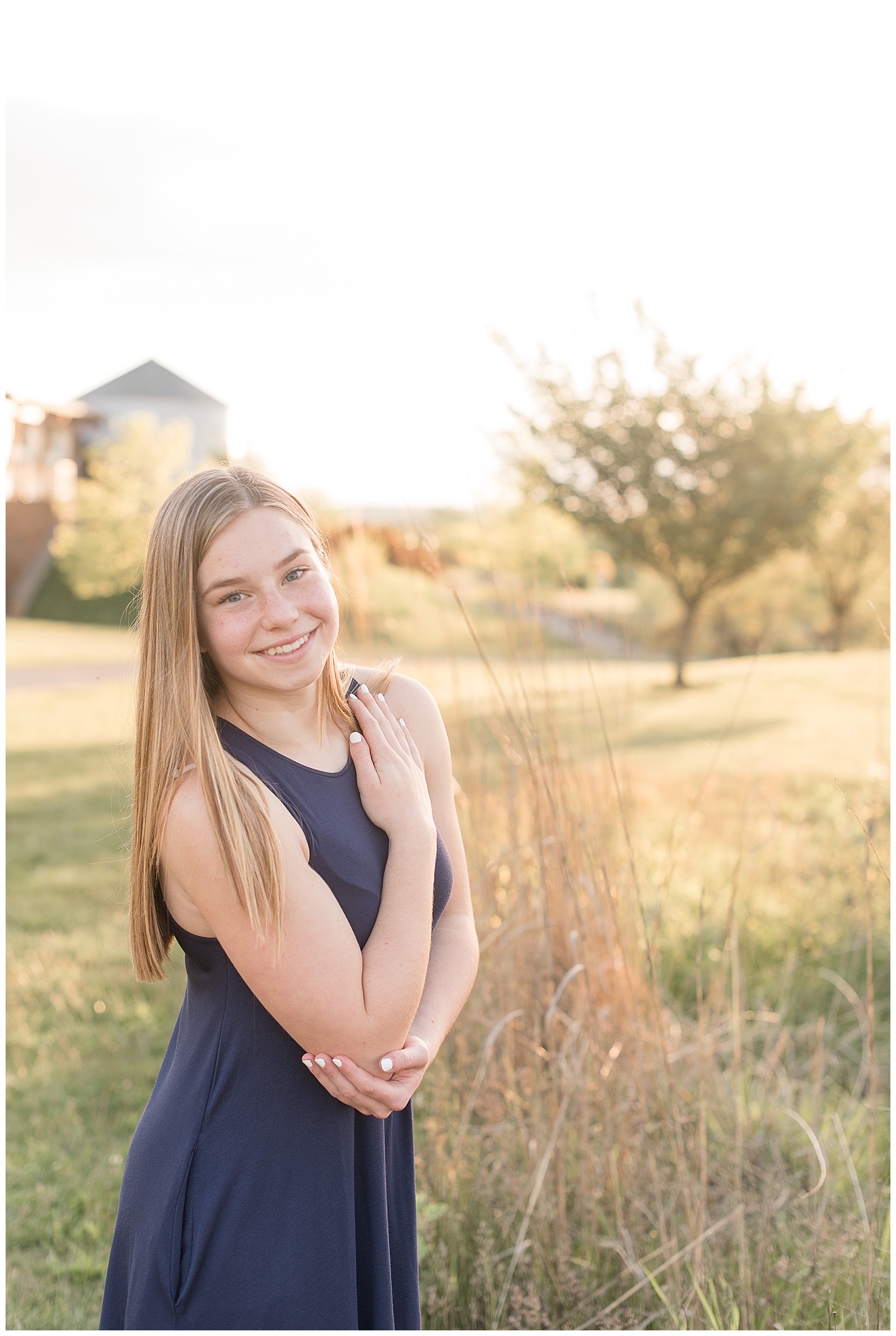 senior girl with right arm crossed over chest smiling at camera with grain bin and tall grasses behind her in lancaster pennsylvania