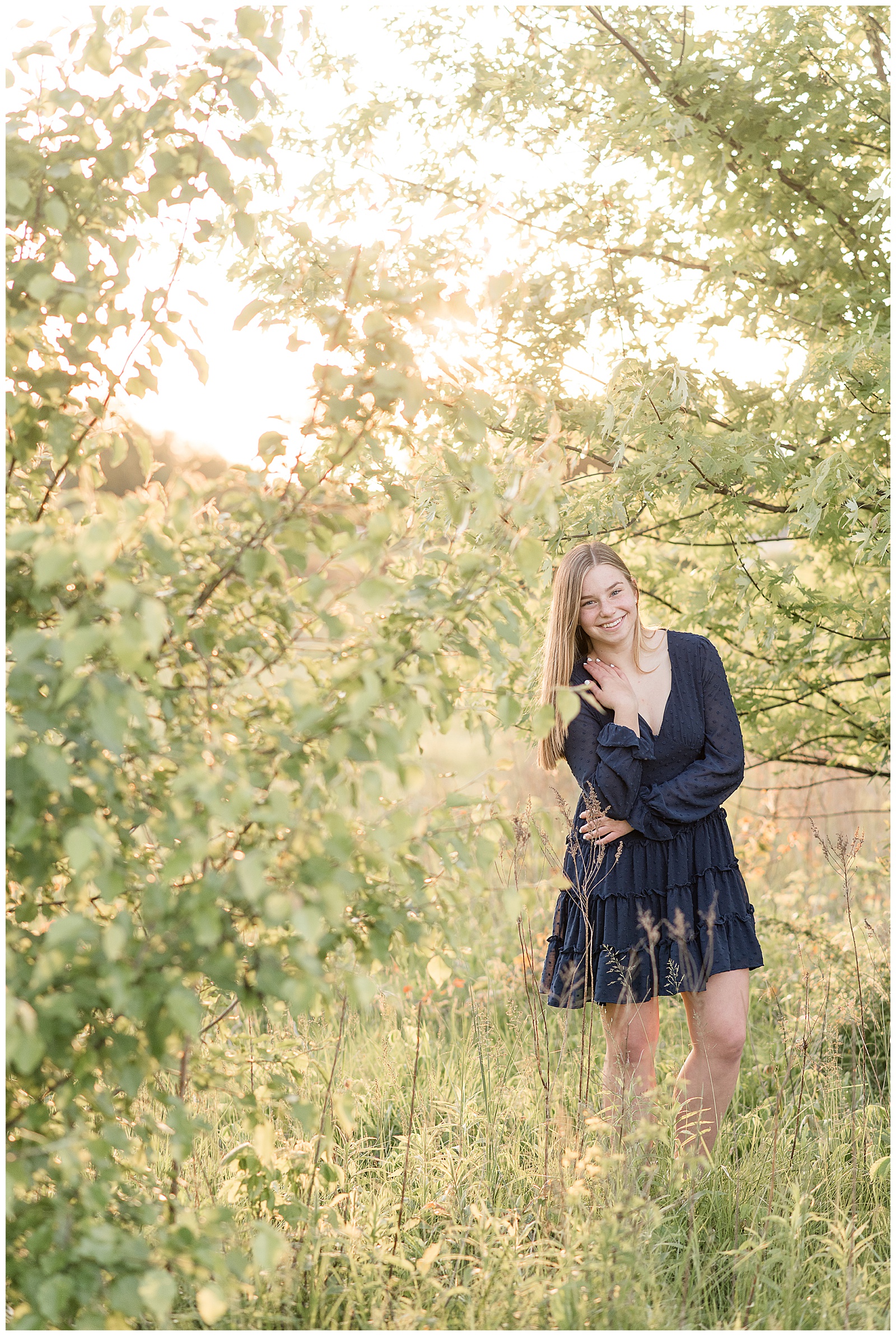 senior girl standing near tree branches with bright sun shining behind her as she smiles at camera at overlook park