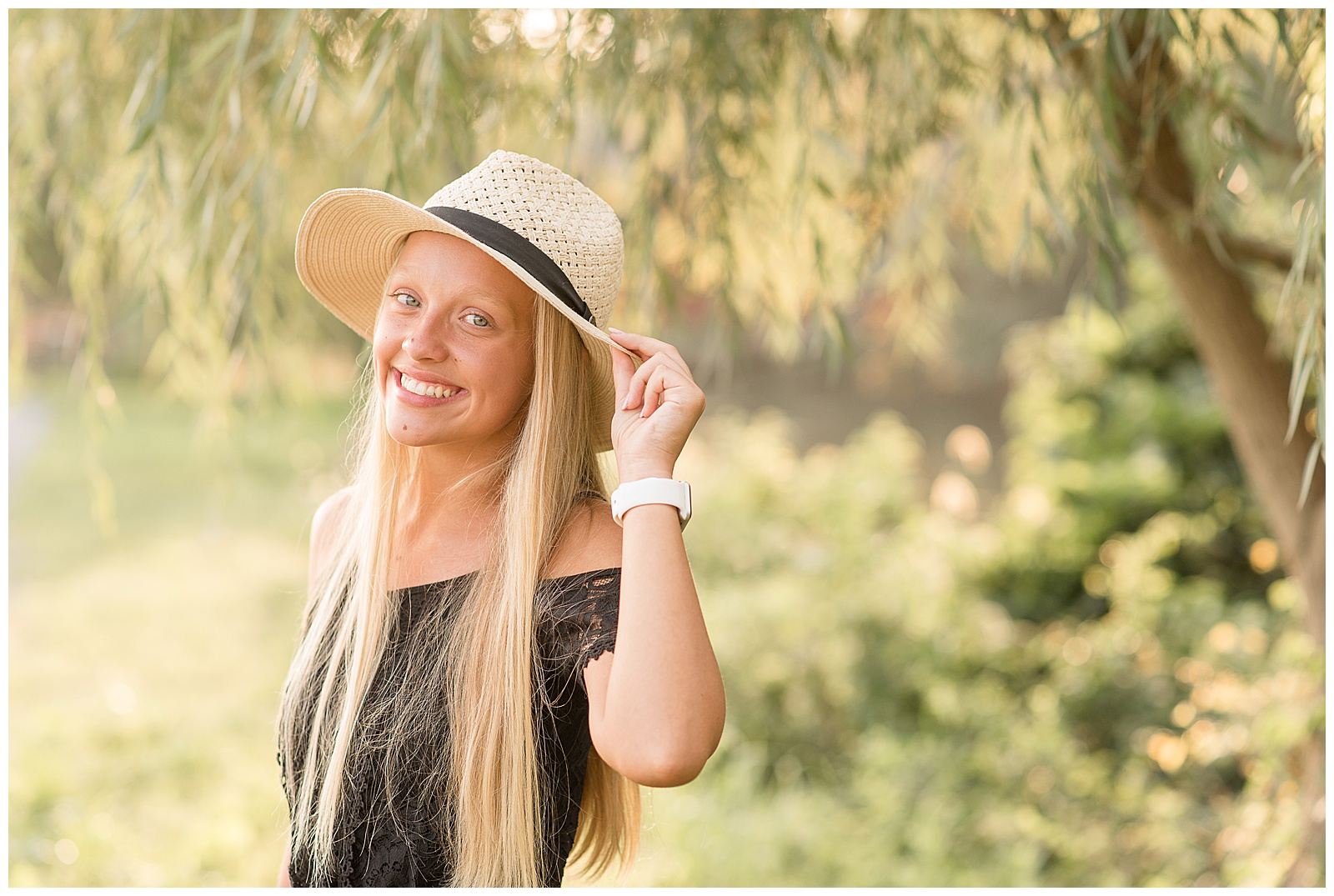 senior girl holds brim of her hat with left hand as she smiles at camera on sunny evening at messiah college