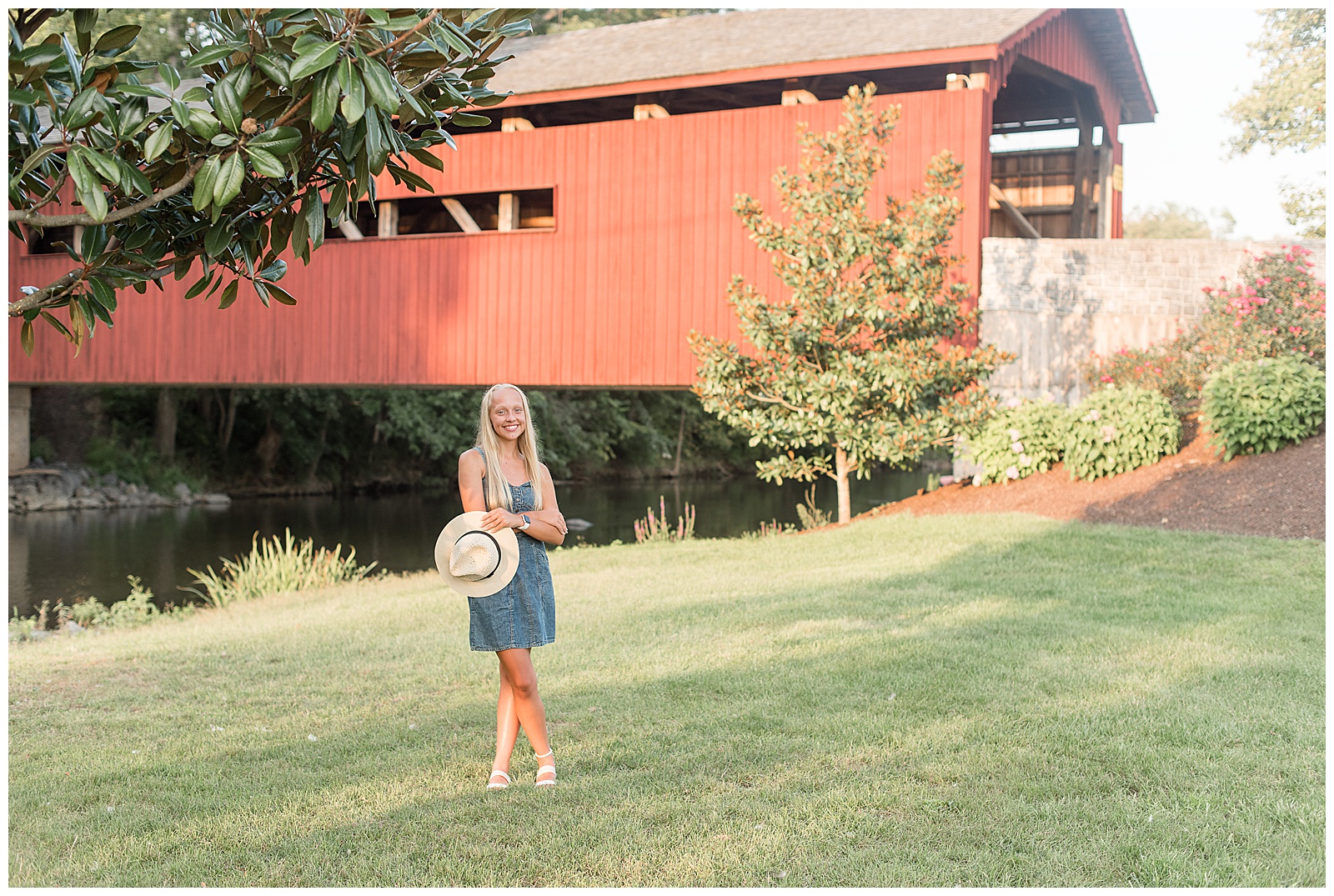 senior girl holds hat in left hand and smiles by red covered bridge on sunny evening at messiah college