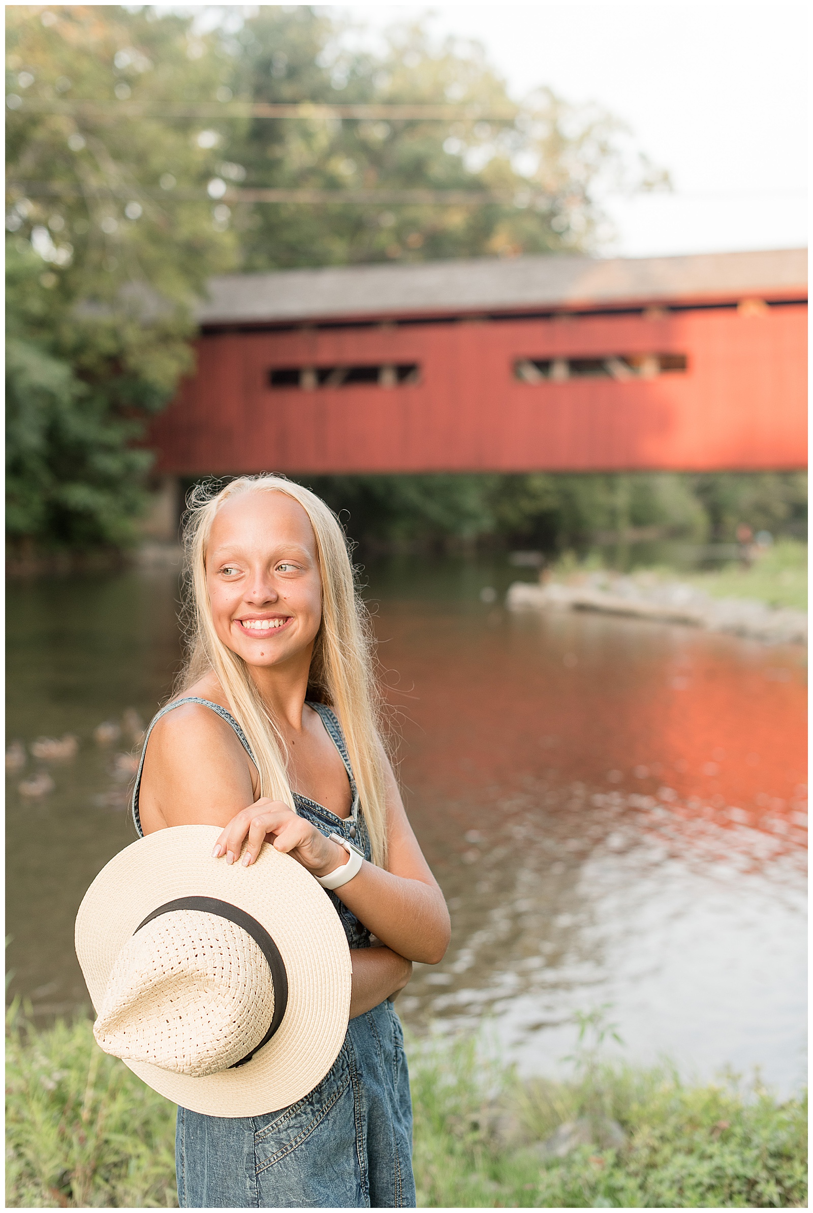 senior girl looks over right shoulder and holds trendy hat in left hand with red bridge behind her at messiah university