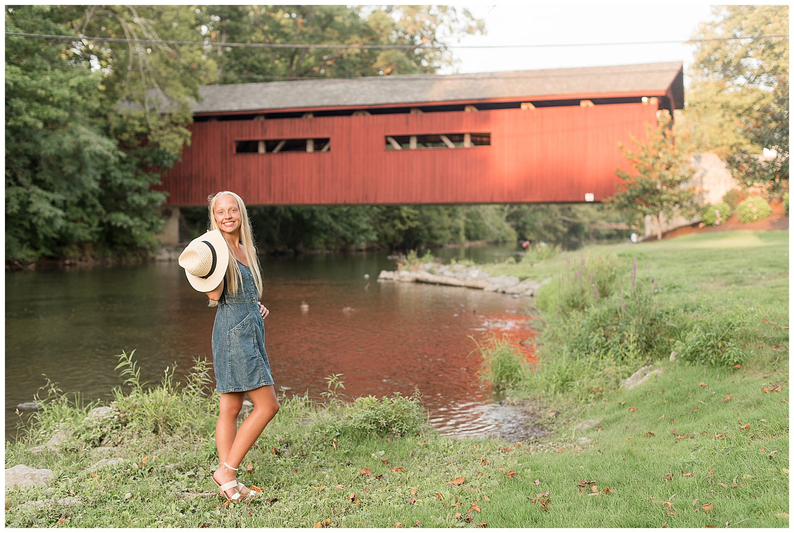 senior girl stands along creek with red covered bridge behind her on sunny summer evening in cumberland county pennsylvania