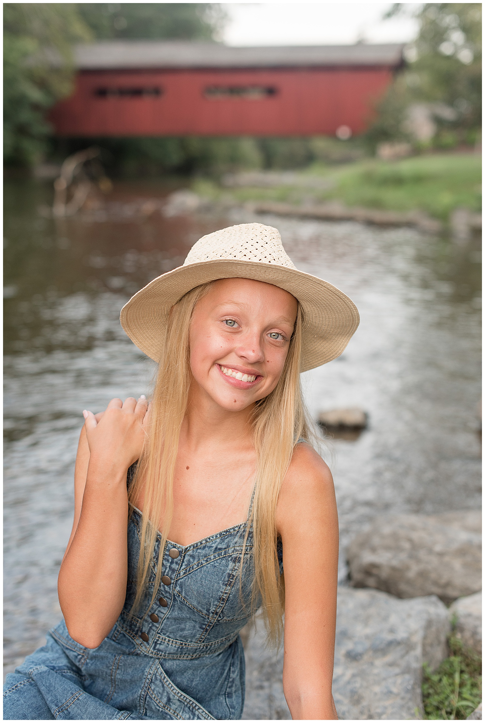 closeup photo of senior girl sitting on rocks by creek with right hand on right shoulder and red covered bridge behind her in pennslvania