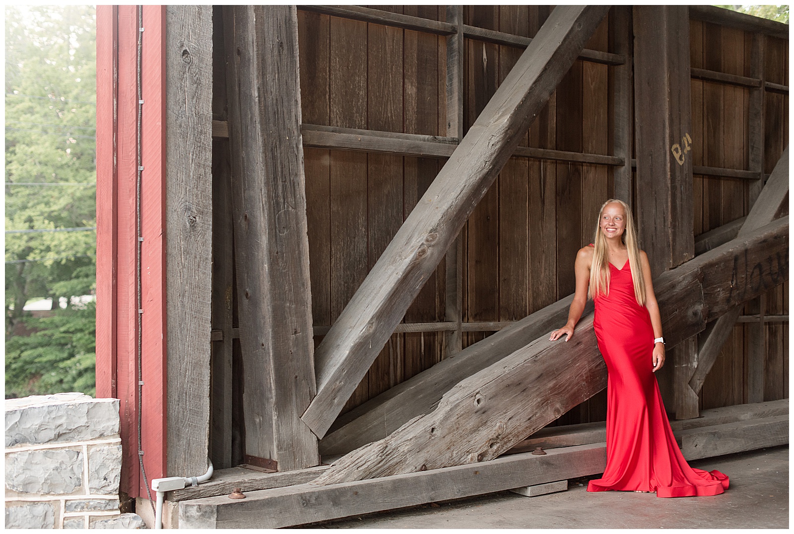 senior girl in long red dress leans right hand on wooden beam of covered bridge and smiles on sunny evening at messiah university