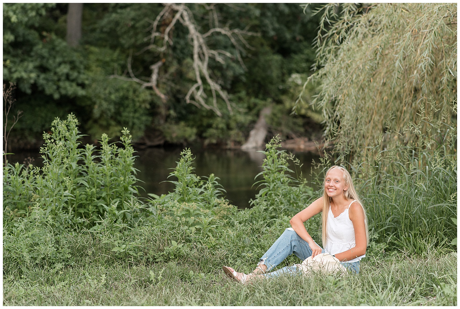 senior girl wearing white tank top and blue jeans sits with right elbow on right bent knee by creek at messiah university