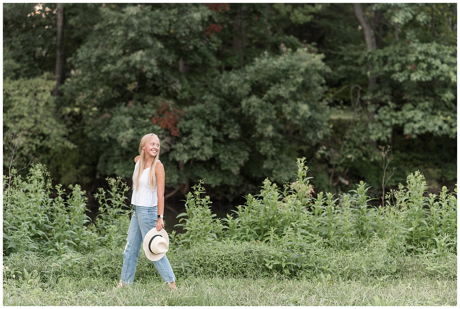 senior girl in white tank top and blue jeans holds hat in left hand and walks look over left shoulder by stream in pennsylvania