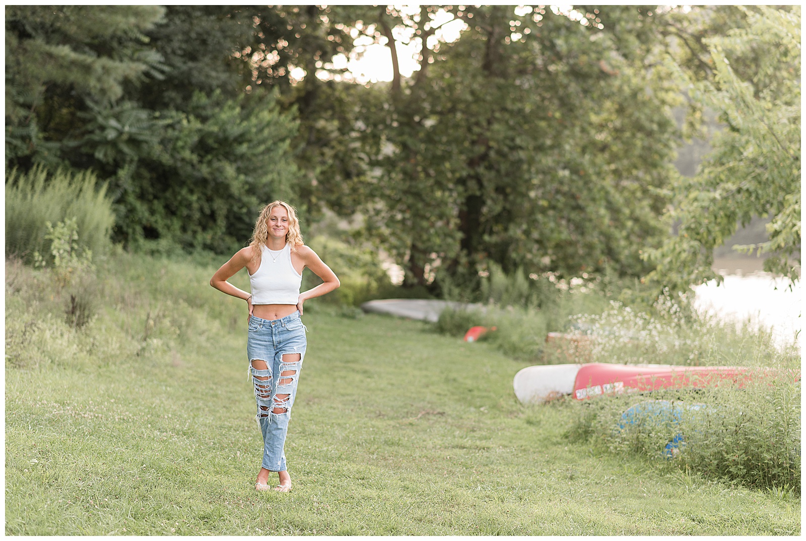 senior girl standing in grass by row of colorful canoes with hands on hips at speedwell forge lake