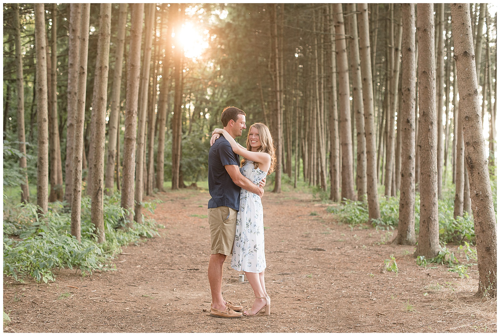 couple standing close in pine tree grove at overlook park with sun peeking through trees behind them on summer night