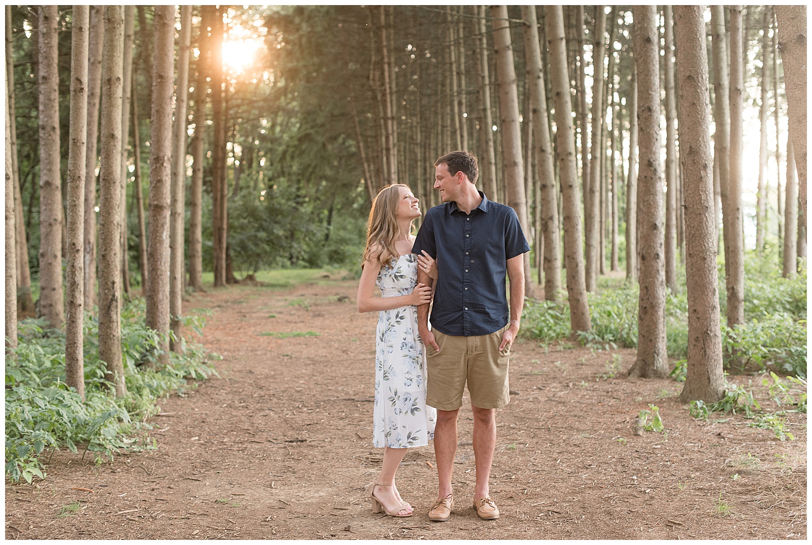 engaged couple looking at each other smiling in pine tree grove with girl in floral dress and guy in button up shirt and khaki shorts
