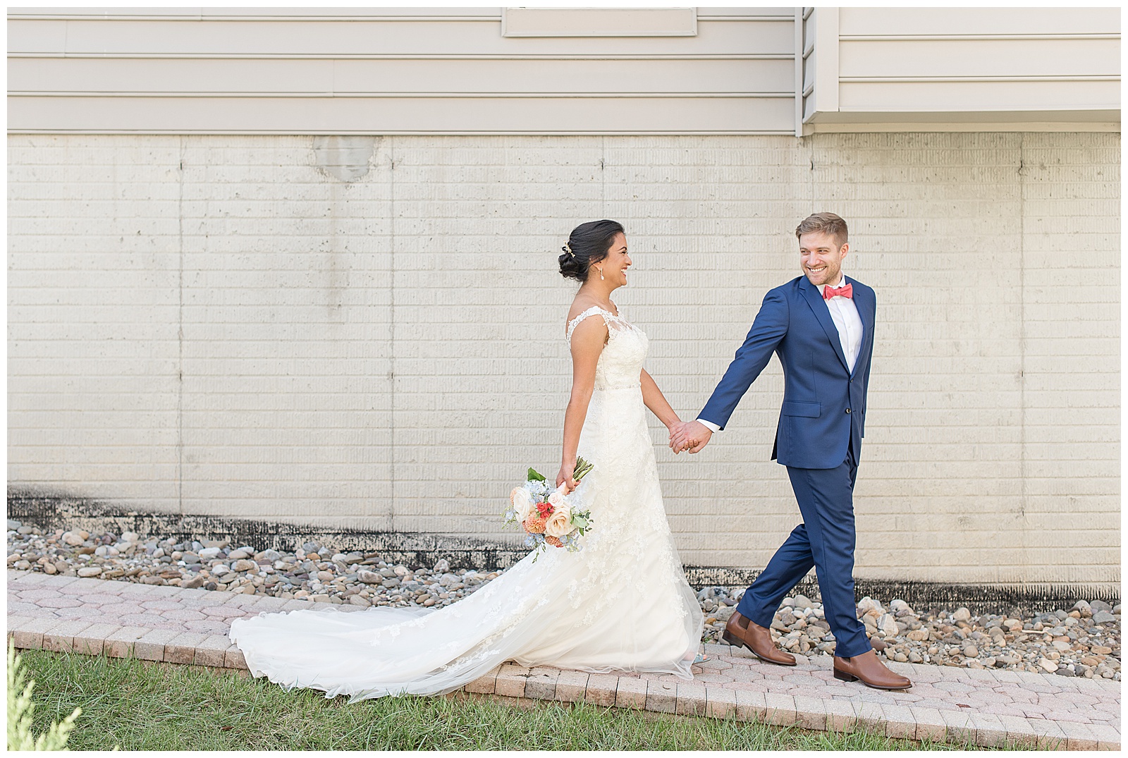 groom holds bride's left hand leading her down pathway along building on sunny day in maryland