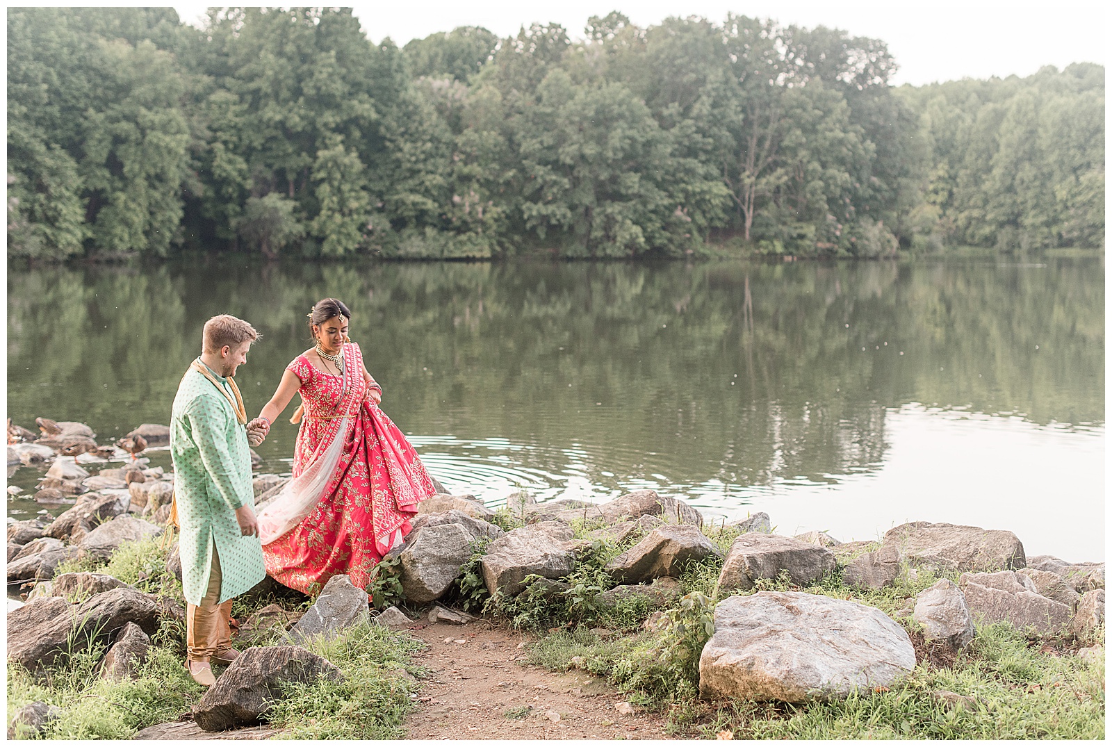 groom leading his bride off of large rocks by pond as they wear their traditional indian wedding outfits in ellicott city maryland
