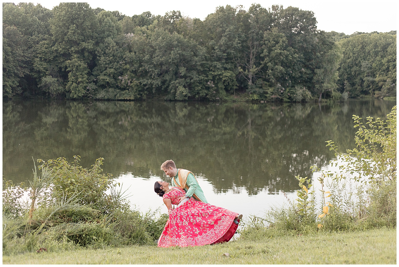 groom dips his bride back beside pond in maryland as they both wear tradition indian wedding clothes