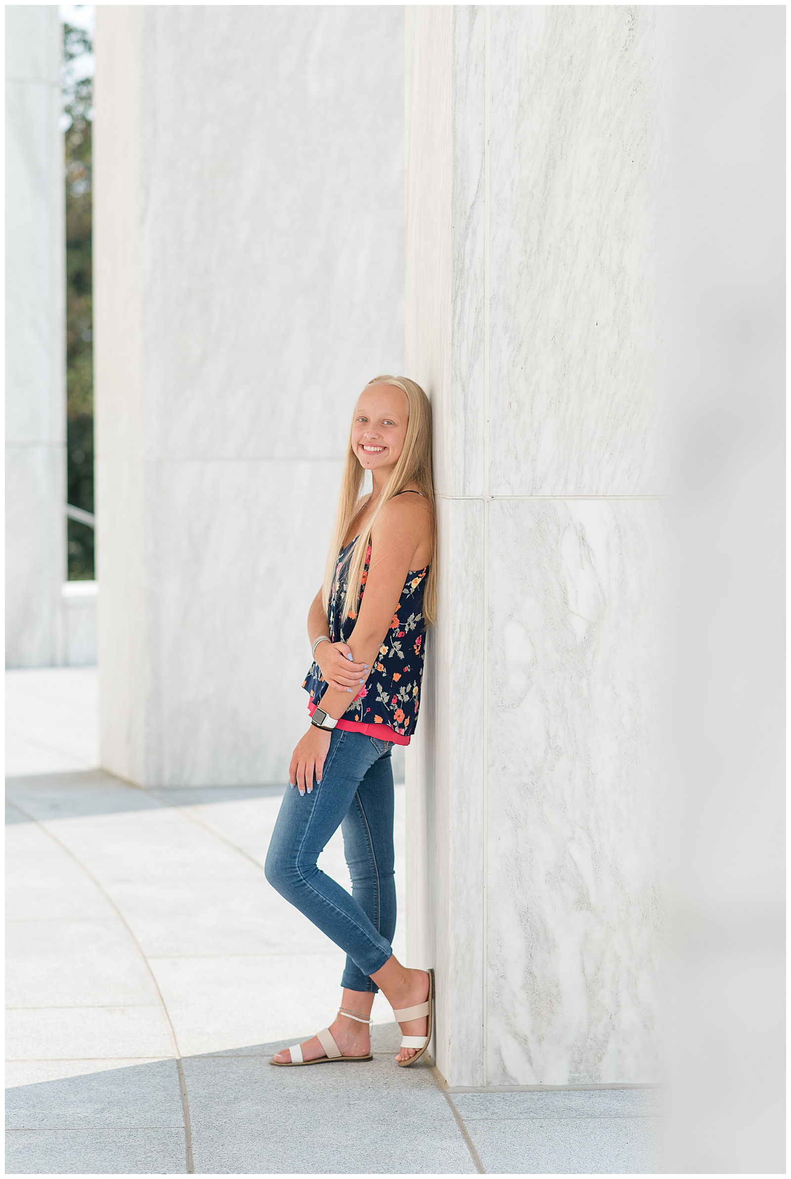 senior girl in floral tank top and blue jeans leaning her back against white wall at founders hall in hershey pennsylvania