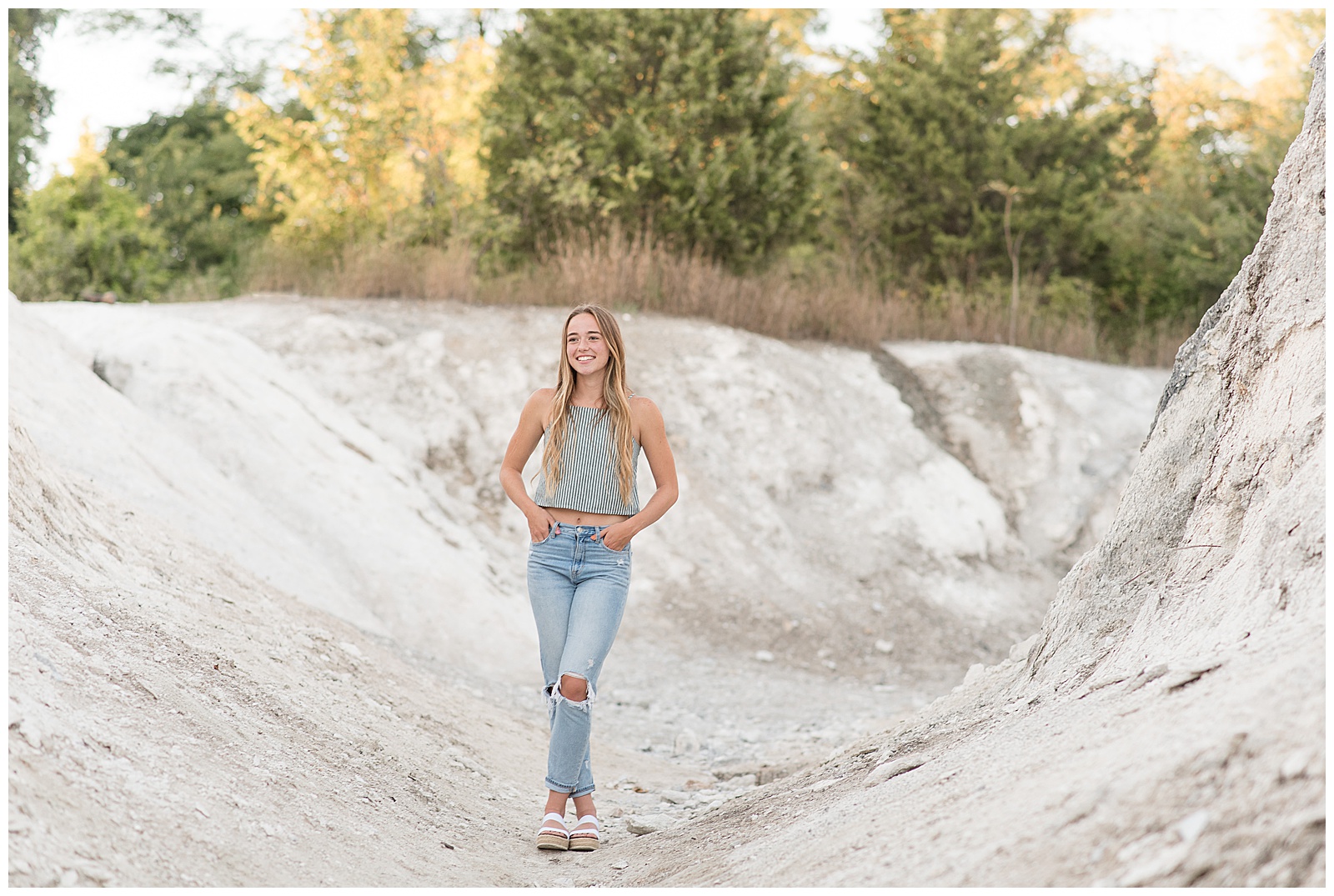 senior girl with hands in jean pockets and left ankle crossed over right ankle standing on white cliff with trees behind her in lancaster pennsylvania