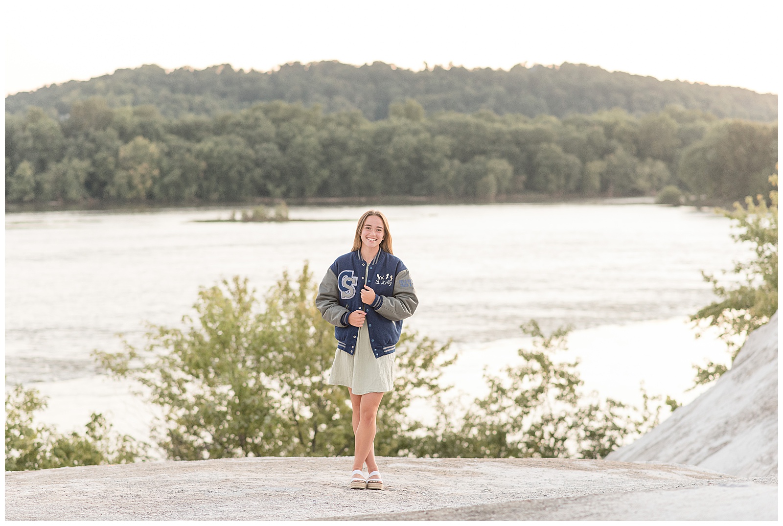 senior girl standing atop white cliffs of canoy with susquehanna river behind her as she wears her varity jacket overtop her summer dress