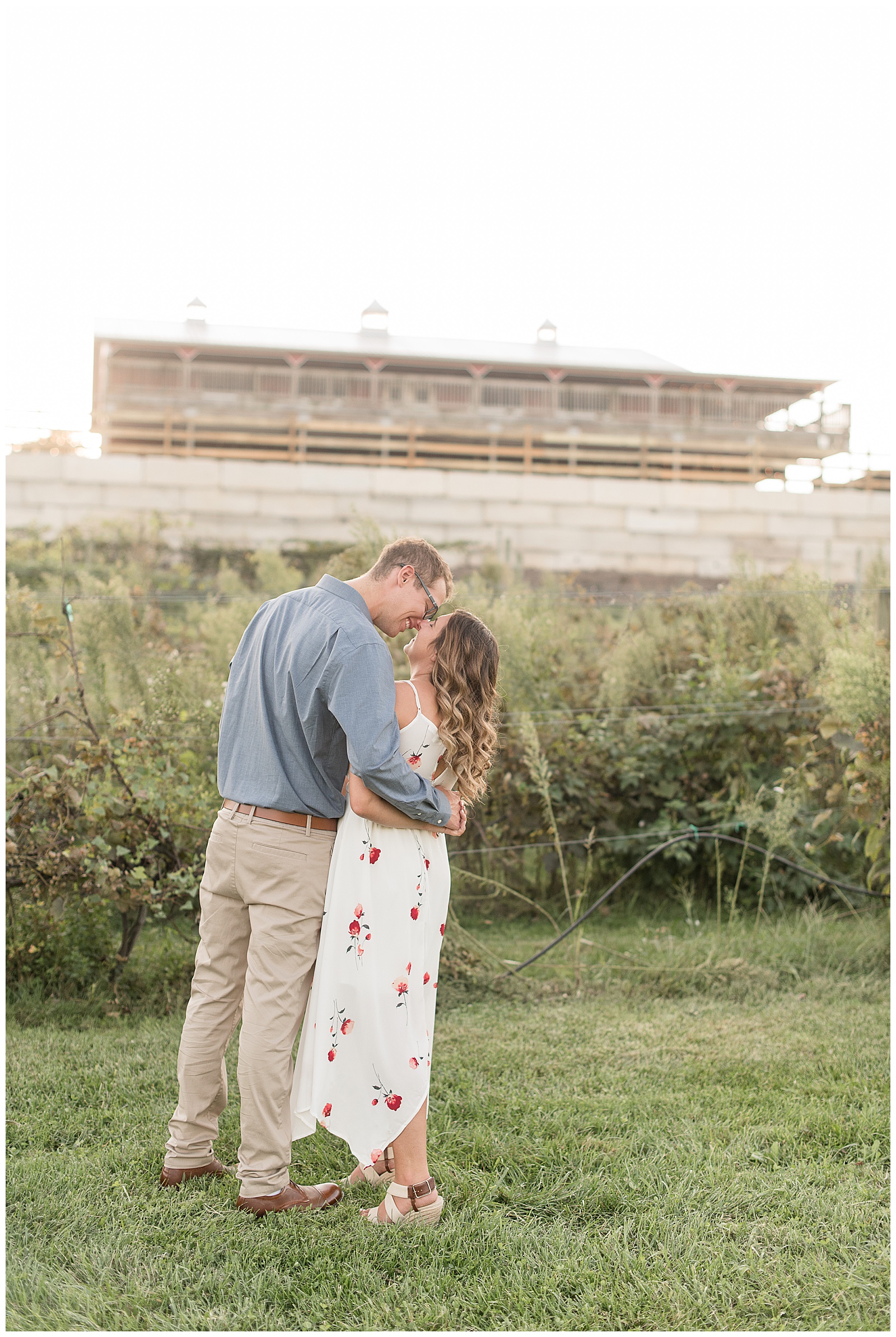 engaged couple with their backs toward camera kissing with vineyards and large white building in the background in hershey pennsylvania