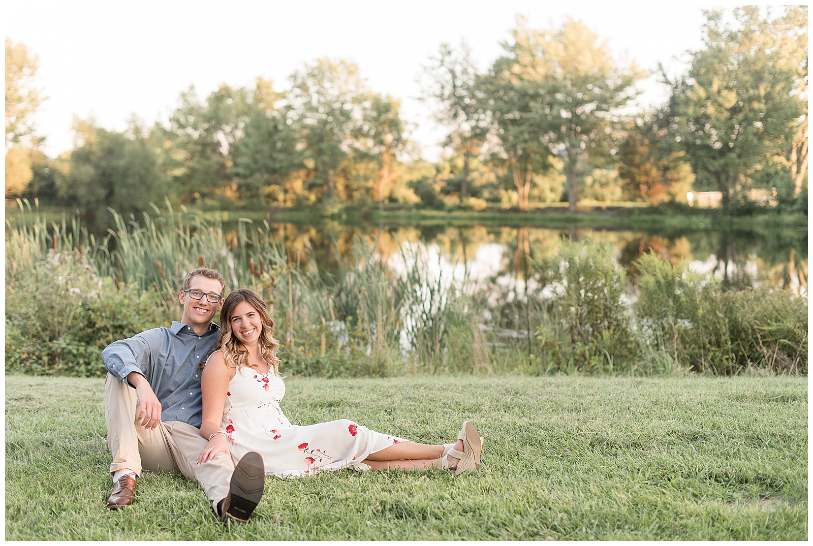 engaged couple sitting in the grass with girl leaning back against guy and pond behind them at vineyards at hershey on sunny evening