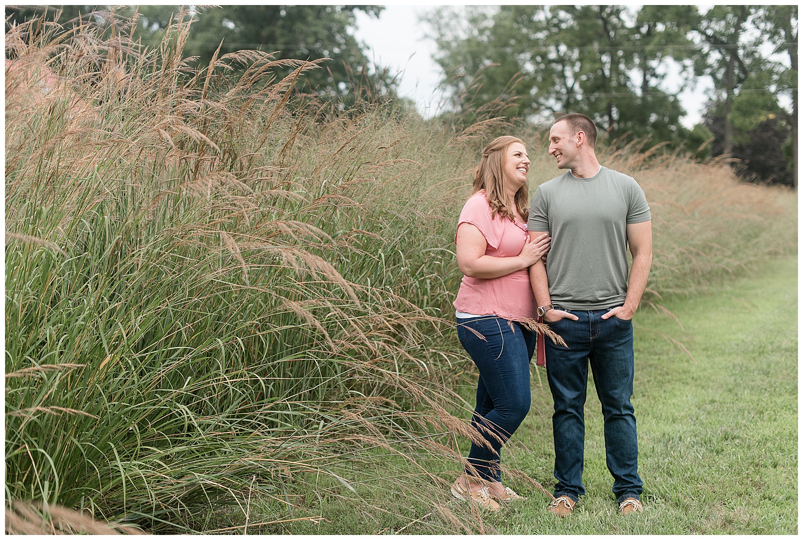engaged couple standing close looking at each other smiling beside tall wild grasses on cloudy day at hibernia park in coatesville pennsylvania
