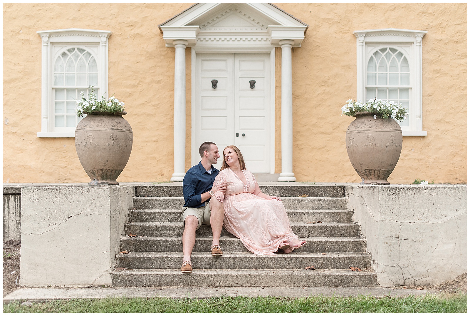 engaged couple sitting on concrete steps of yellow brick house smiling and looking at each other at hibernia county park