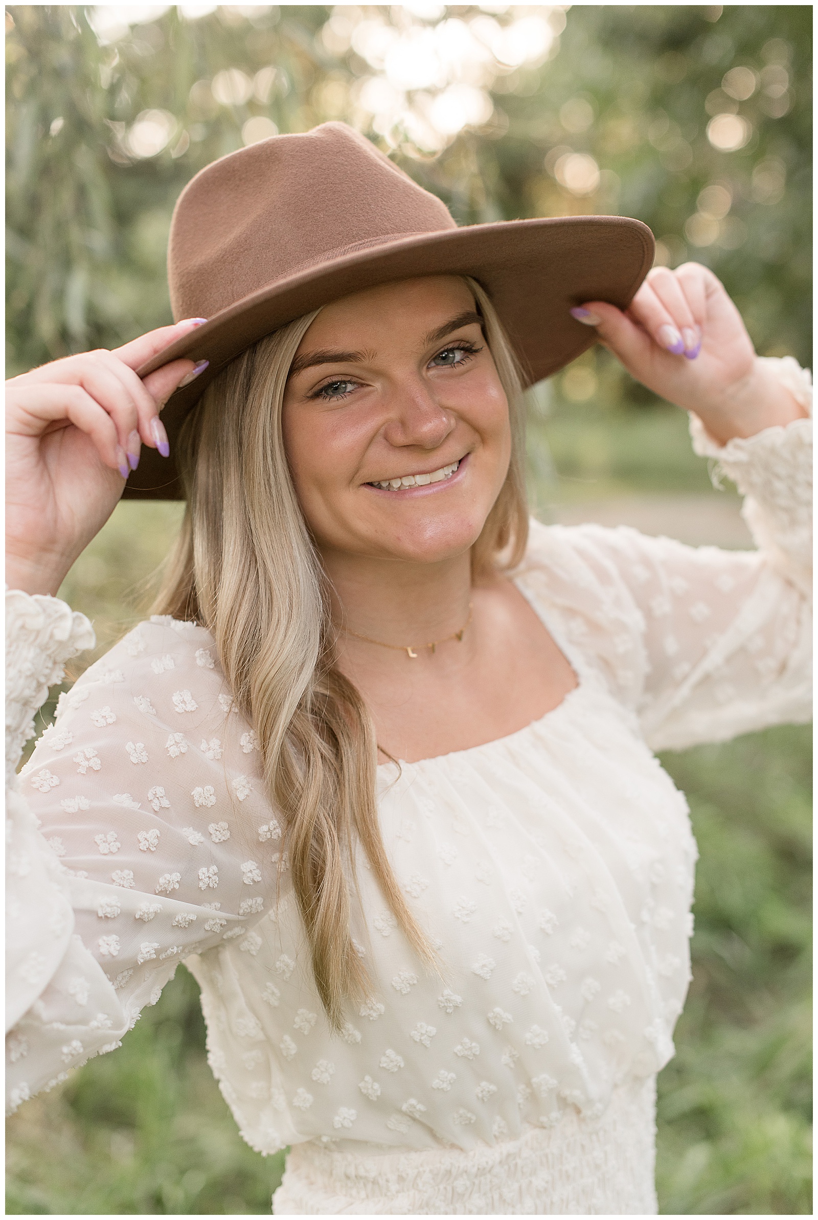 senior girl wearing ivory dress and tan hat holding the brim of hat with each hand and smiling at overlook park