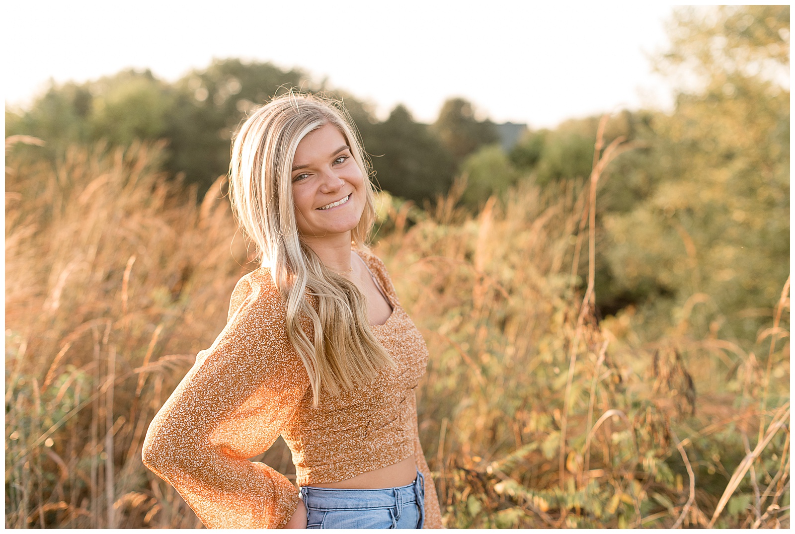 senior girl with right should towards camera and right hand on hip smiling with sunlight behind her near tall grasses in lancaster pennsylvania
