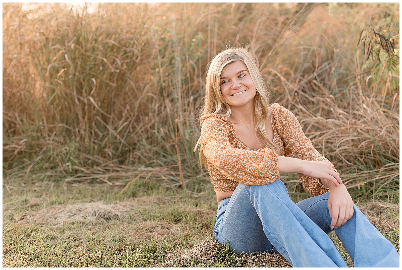 senior girl sitting by tall wild grasses smiling looking off towards her right shoulder with her elbows resting on her bent knees at overlook park