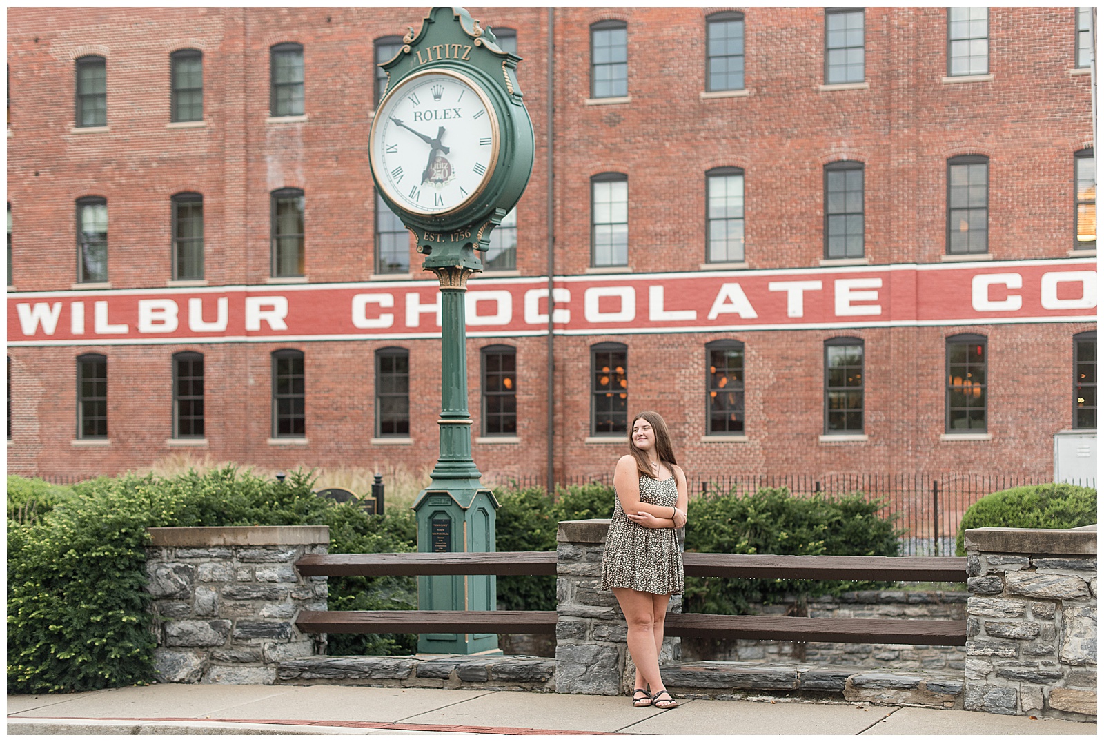 senior girl in flowy dress standing by old clock outside historic wilbur chocolate factory building in lititz pennsylvania