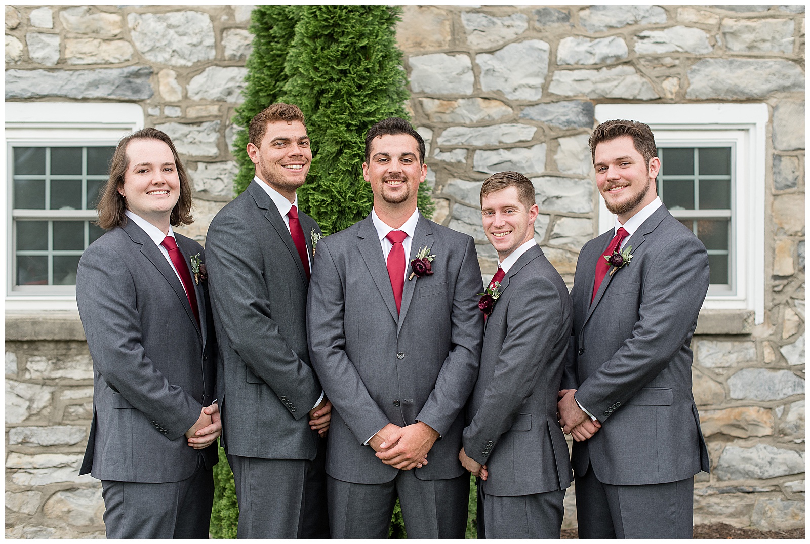 groom with his four groomsmen all wearing dark gray suits with red ties and their hands folded in front of them by stone wall at the barn at silverstone