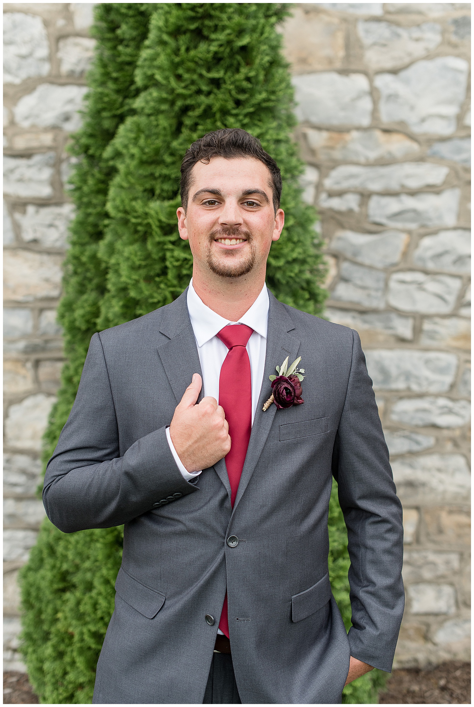 groom smiling at camera with right hand holding edge of his gray suit with red tie at the barn at silverstone
