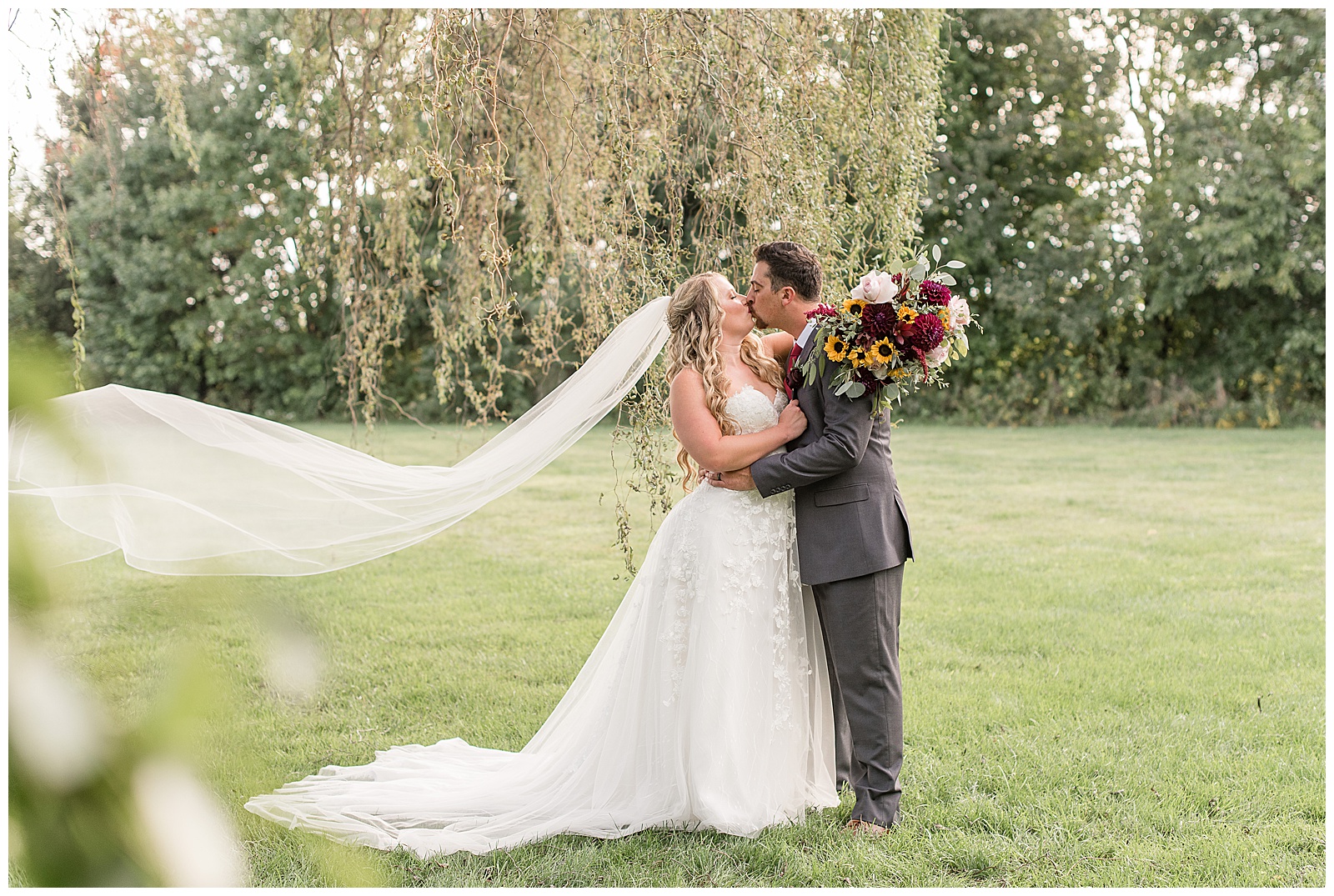 couple kissing as she holds her bouquet around groom's neck and her long veil blows in the breeze behind her at the barn at silverstone