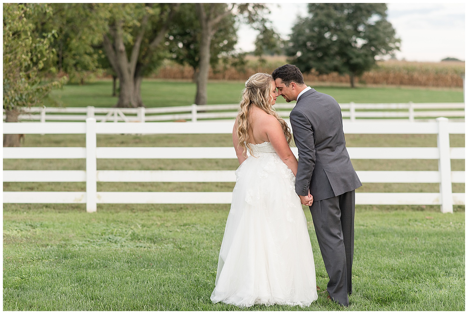 couple's backs toward camera as their foreheads touch and they hold hands while standing on lawn by white fence at the barn at silverstone