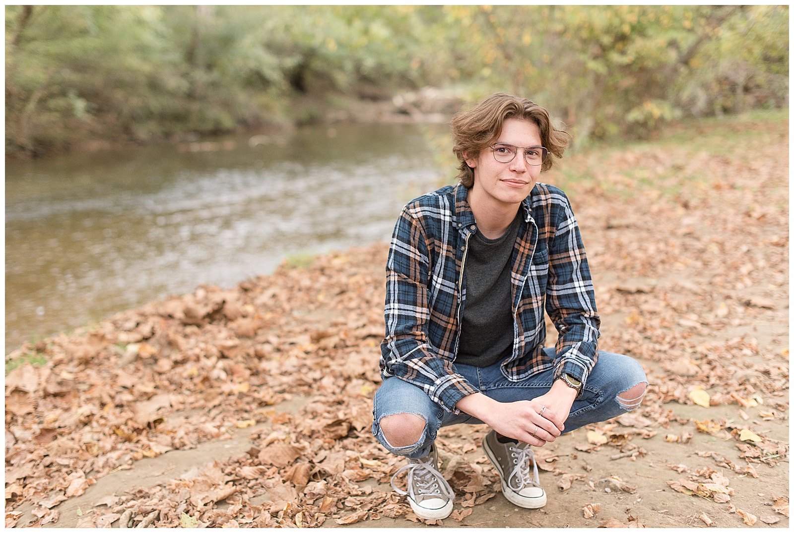 male senior spokesmodel crouching down with elbows on knees and hands together on dry leaves by river at lancaster county park on fall day