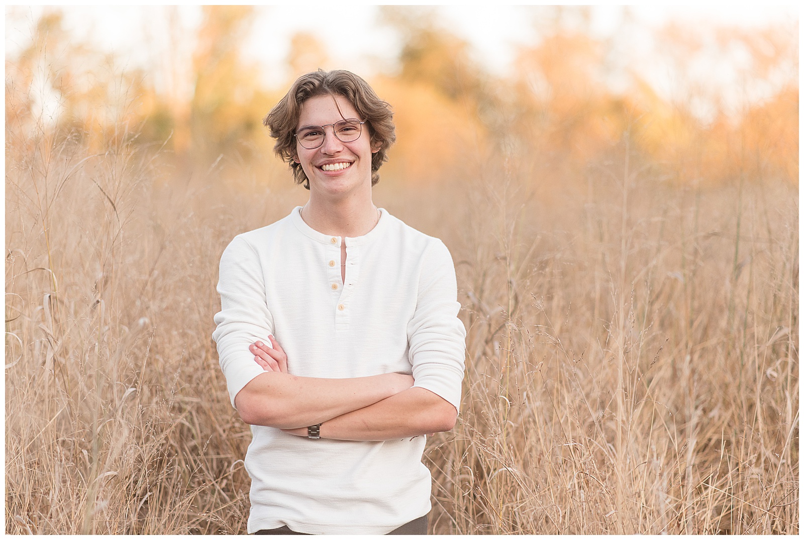 senior guy wearing white henley shirt and blue jeans with arms folded smiling at camera with dried out wild grasses surrounding him at lancaster county park