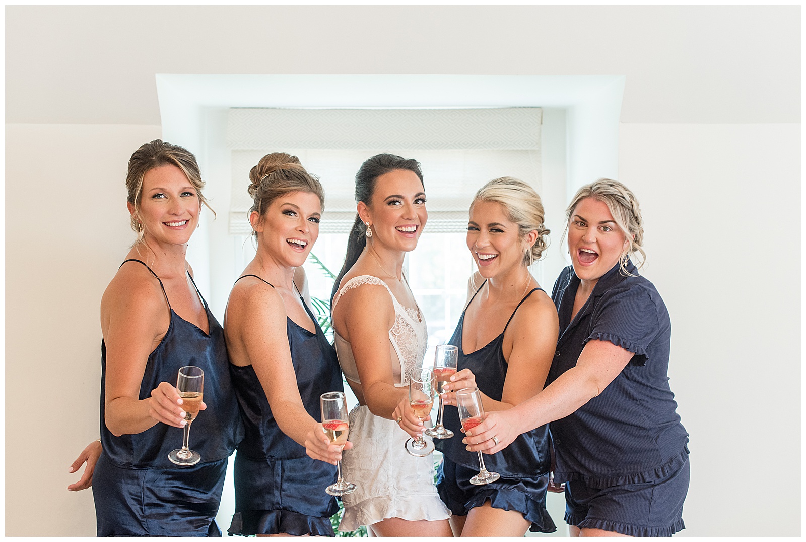 bride in white slip surrounded by her four bridesmaids in navy blue silky pajamas toasting in bridal suite in north jersey country club