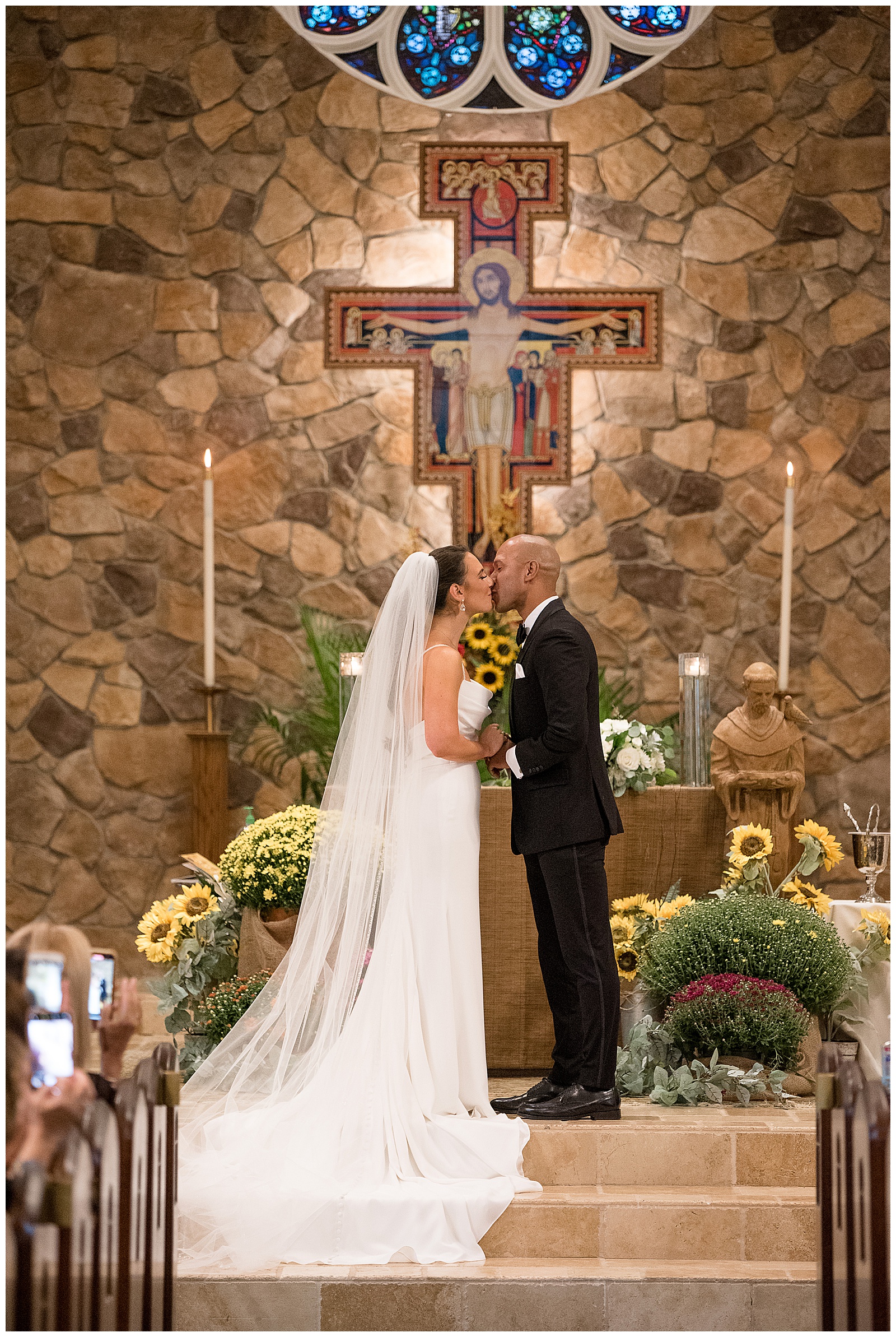 bride and groom kissing during wedding ceremony inside catholic church with beautiful cross and stone wall behind them in new jersey