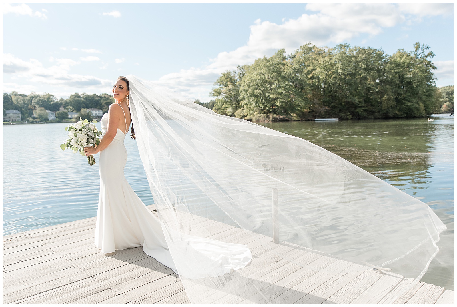 bride looking back over left shoulder at camera with long veil blowing behind her as she stands on dock with water surrounding her in new jersey