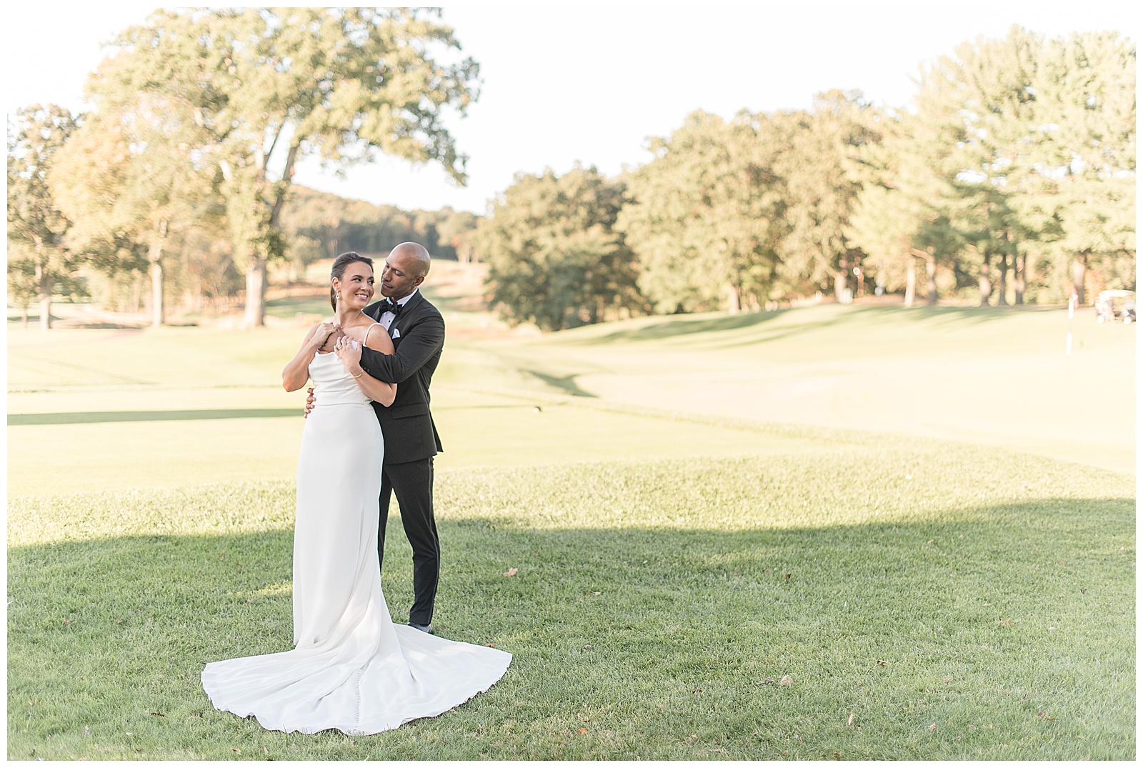 groom hugging bride from behind as she holds onto his arms and they smile on sunny golf course at north jersey country club